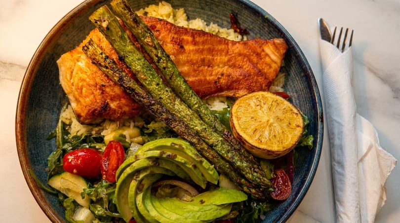 Grilled salmon with asparagus, avocado, cherry tomatoes, and lemon over rice in a blue bowl.